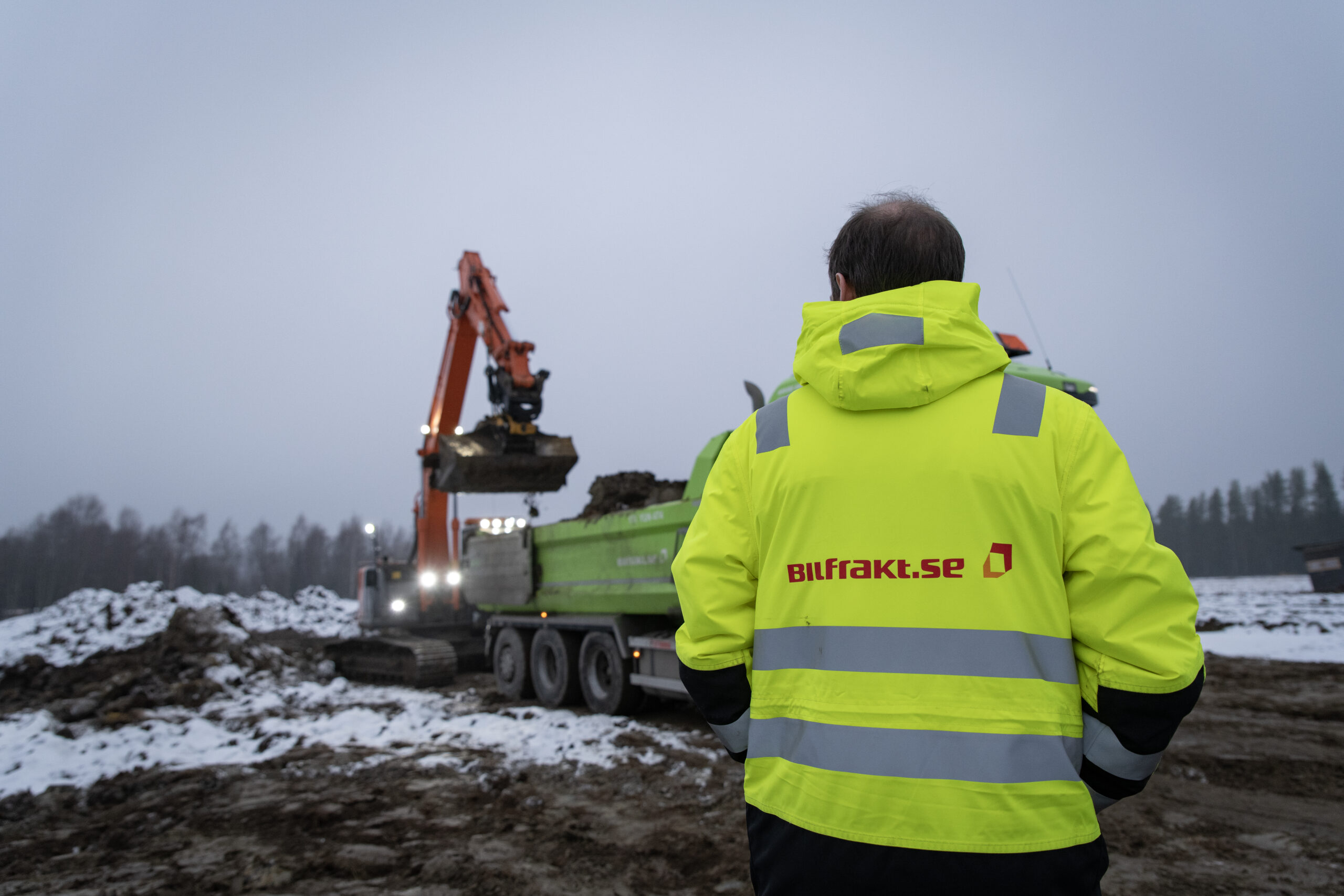 En person i varseljacka från Bilfrakt övervakar lastningsarbetet på en byggarbetsplats